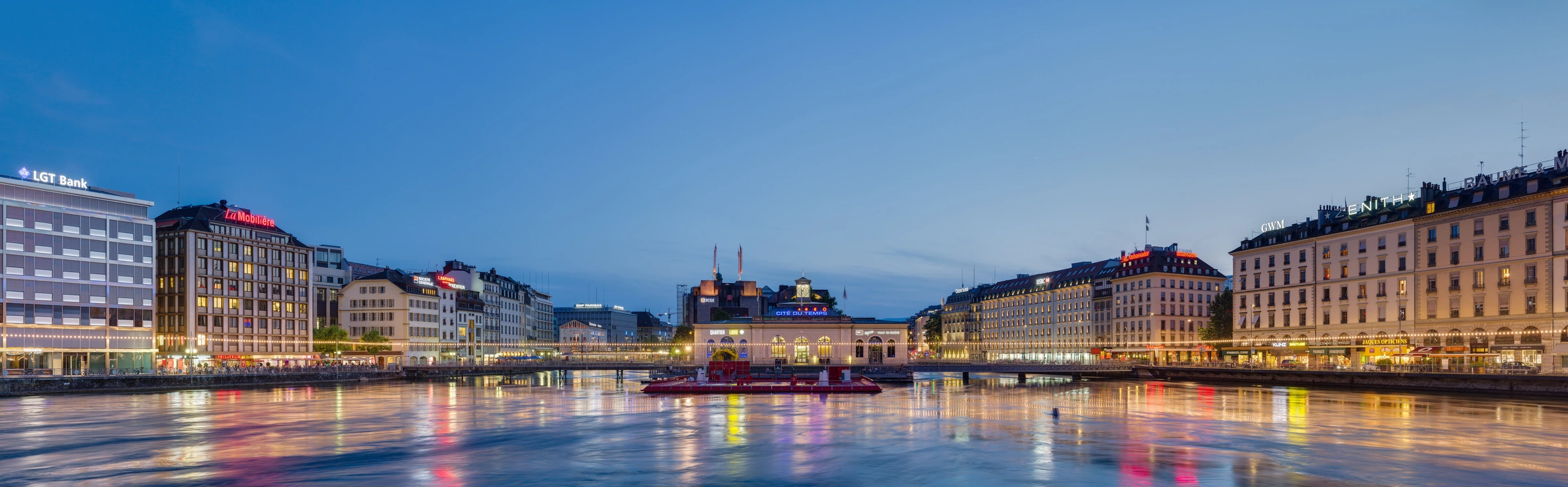 Geneva at blue hour, with the Pont de la Machine, illuminated waterfront buildings and reflections across the Rhône.