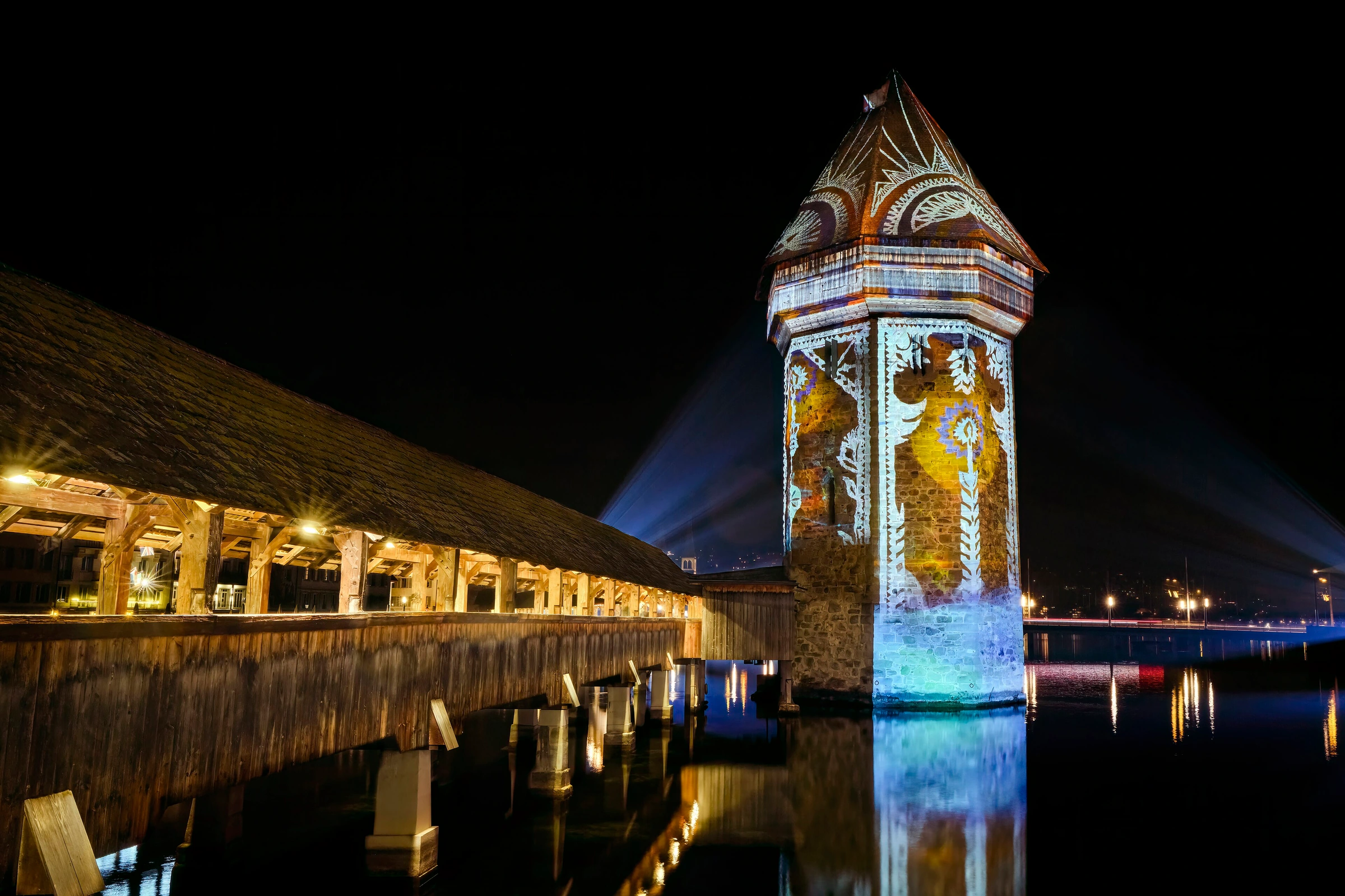 Chapel Bridge and the Water Tower reflected in the Reuss at night during the Lilu Light Festival in January 2025. The patterns on the tower are real festival light projections.