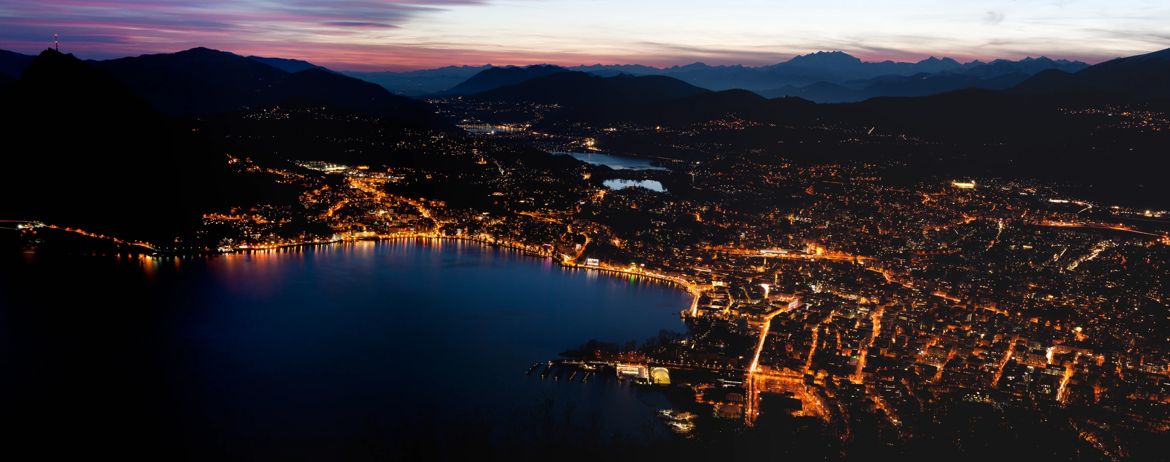 Lugano at twilight from Monte Brè, with pink sunset clouds and warm city lights reflected in the lake.